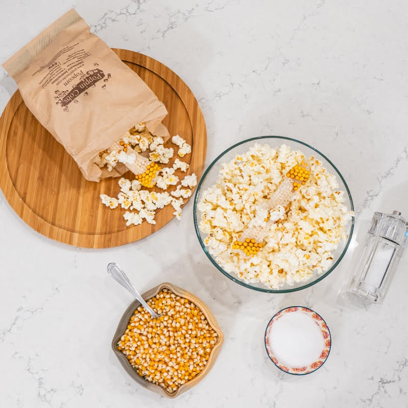 A collection of popcorn cobs on a wooden plate, with a glass bowl filled with popcorn, a shaker of salt, and a brown bag labeled 'Poppin Cobs' in the background.