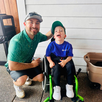 Man kneeling smiling next to a little boy in a green wheelchair 