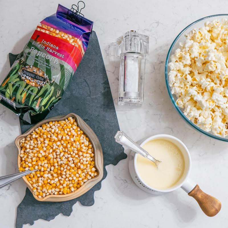 A bowl of popcorn kernels on a dark surface, a package of popcorn kernels in the background, a mug in front, and a wooden spoon to the side, all on a light background.